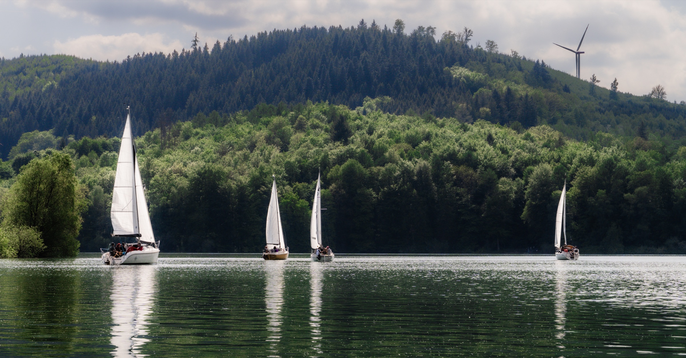 Weites Panorama des Hennesees mit Segelbooten, dahinter ein Windrad auf der Kuppe