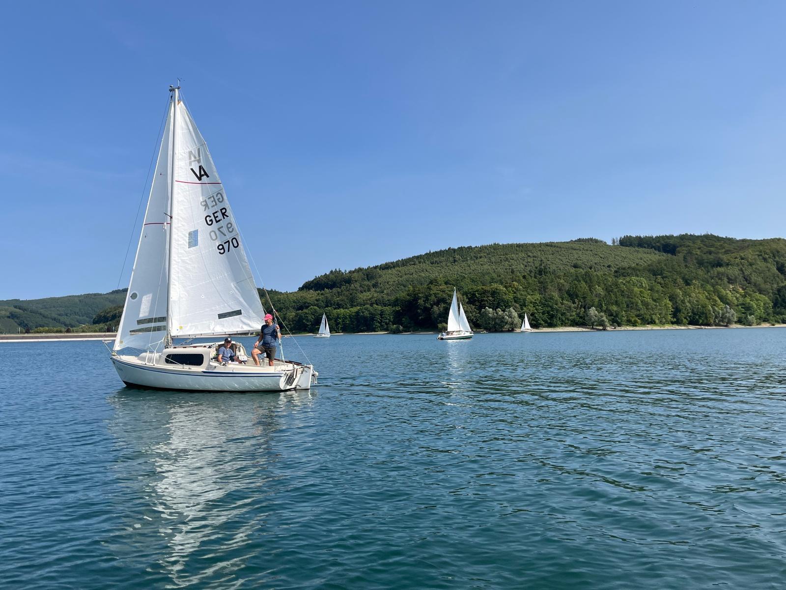 Segelboot GER 970 mit zwei Seglern auf dem sommerlich blauen Hennesee, dahinter bewaldete Hügel und weitere Boote